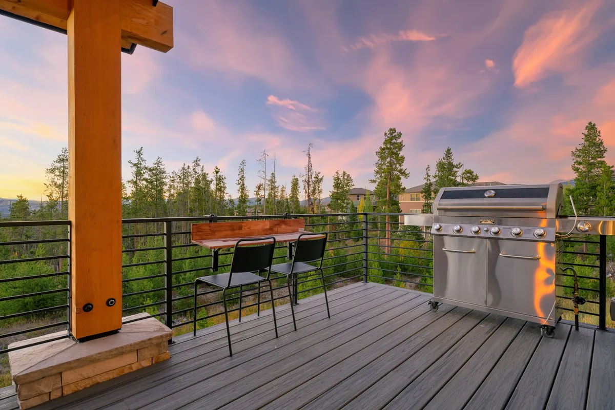 An inviting outdoor grilling and dining area with a modern grill, comfortable outdoor seating, and a scenic view of mountains and trees. The sun is setting, casting a golden glow over the space, suggesting memorable evenings spent outdoors with fresh food and great company.