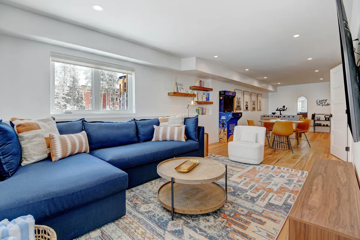 A cozy, casual family room with a large blue sectional sofa, round coffee table, bookshelves filled with books, a television, and an arcade game machine. The space is inviting, with multiple spots for reading, gaming, or relaxing together as a group.
