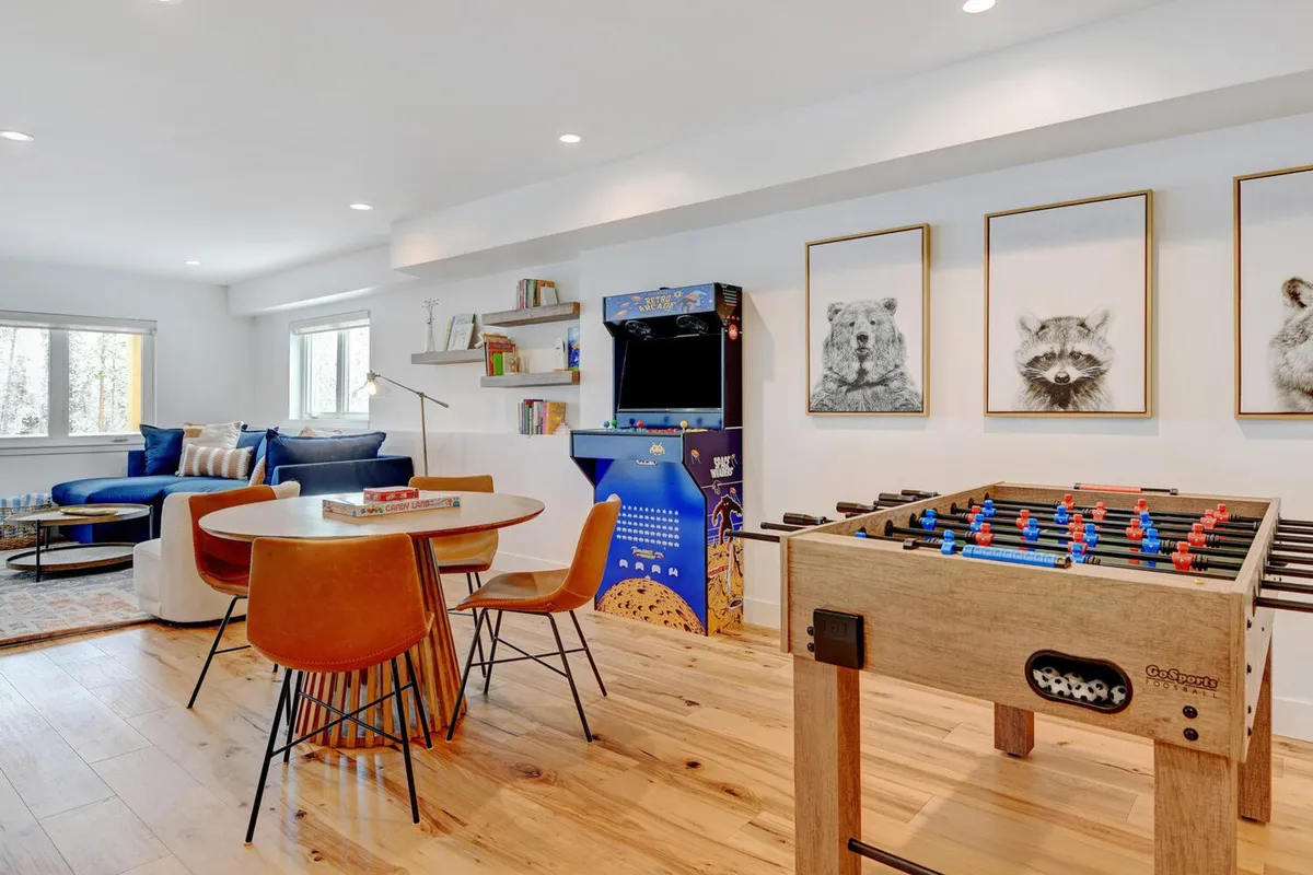 A vibrant interior scene featuring a foosball table and an arcade machine, with a dining table and chairs nearby. Artworks adorn the wall, and the space appears lively and designed for interactive fun and socializing. The presence of different play and gathering options suggests a room that caters to a variety of activities.