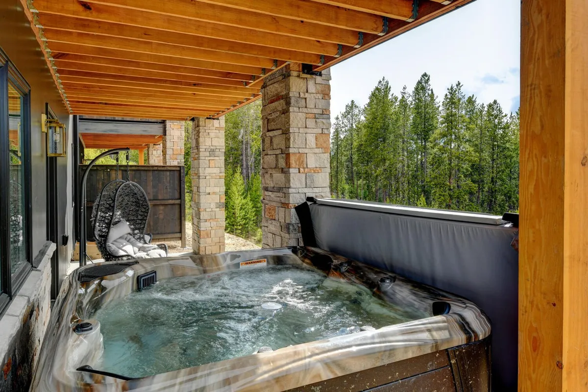 An outdoor hot tub set beneath wooden beams and stone pillars, overlooking a tranquil forest. There's a swing for relaxing and enjoying the fresh mountain air, with the hot tub positioned to maximize views of nature and potential stargazing opportunities.
