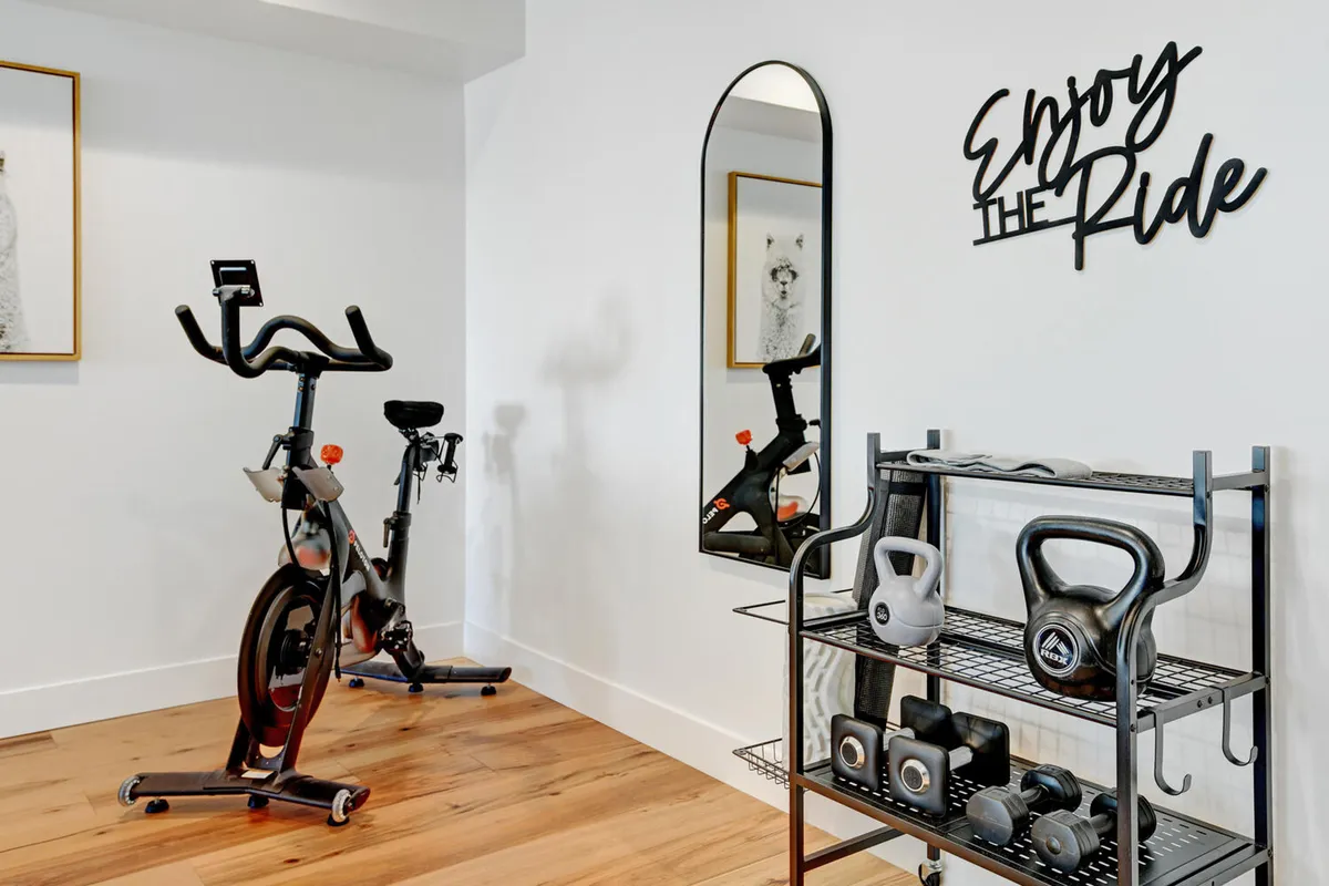 A bright, energetic home gym area featuring an exercise bike, dumbbells, a wall mirror, and decorative motivational wall art. The space is clean and well-lit, ideal for individual or small-group fitness sessions.