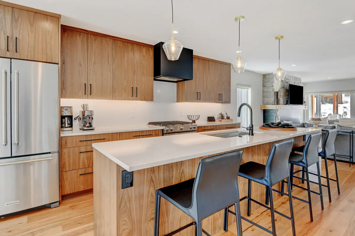 A bright, open kitchen with a spacious island at center, surrounded by barstools. Stainless steel appliances, custom wood cabinetry, and elegant lighting fixtures dominate the space, conveying a modern, chef-ready atmosphere. The open layout hints at adjoining spaces for seamless entertaining.