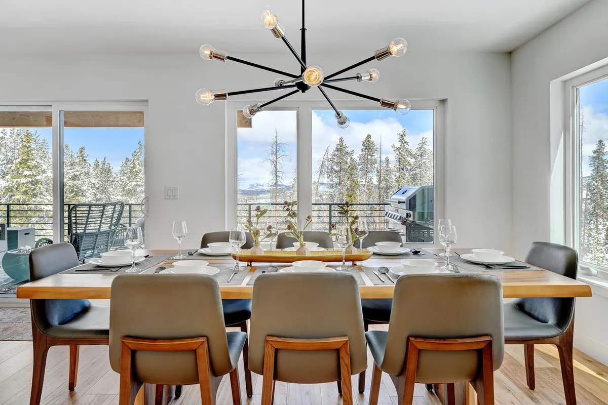 A welcoming dining area features a large table set beneath a stylish chandelier, framed by windows that offer scenic views. The space is ready for group meals, festive gatherings, and socializing, emphasizing warmth and togetherness.