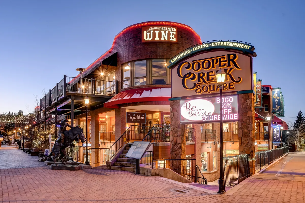 A bustling outdoor plaza with the Cooper Creek Square sign, a wine shop, inviting outdoor seating, and an eye-catching horse sculpture. The atmosphere is vibrant yet welcoming, suggesting a place to enjoy shopping, dining, or gathering with friends and family.
