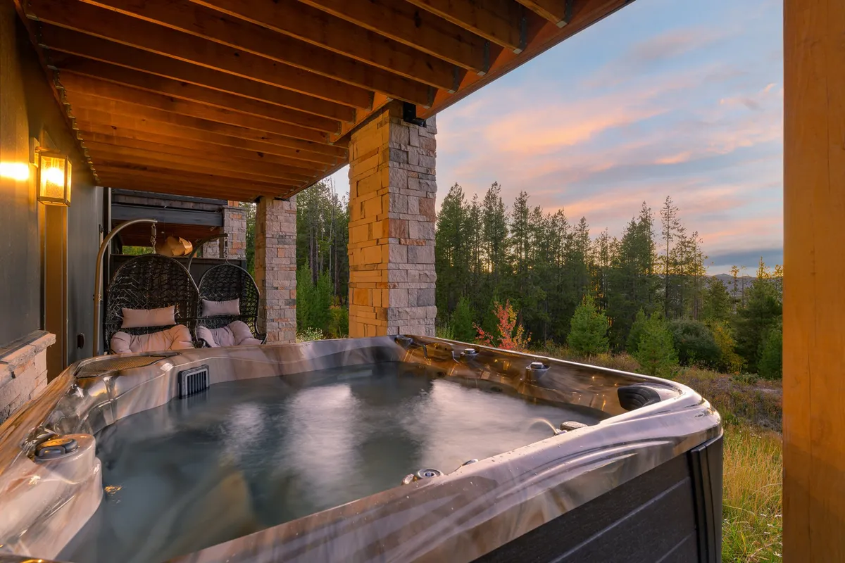 A serene outdoor scene featuring a bubbling hot tub surrounded by tall trees and set beneath a vibrant sunset sky. The hot tub is positioned to maximize the sweeping mountain and forest views, with steam rising gently in the cool air, inviting relaxation in all seasons.