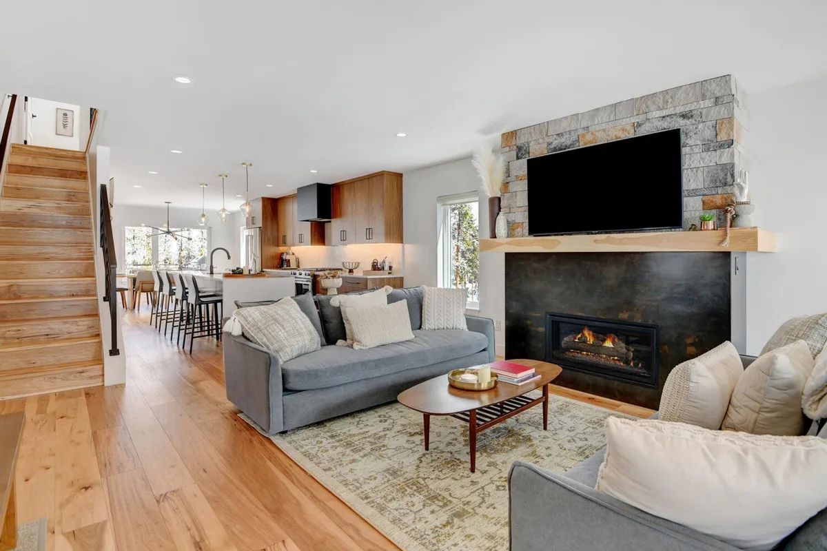 A plush living space with a cozy sofa and coffee table faces a striking fireplace, with the kitchen visible in the background. The mood is comfortable and inviting, perfect for relaxation or unwinding after a day of adventure.